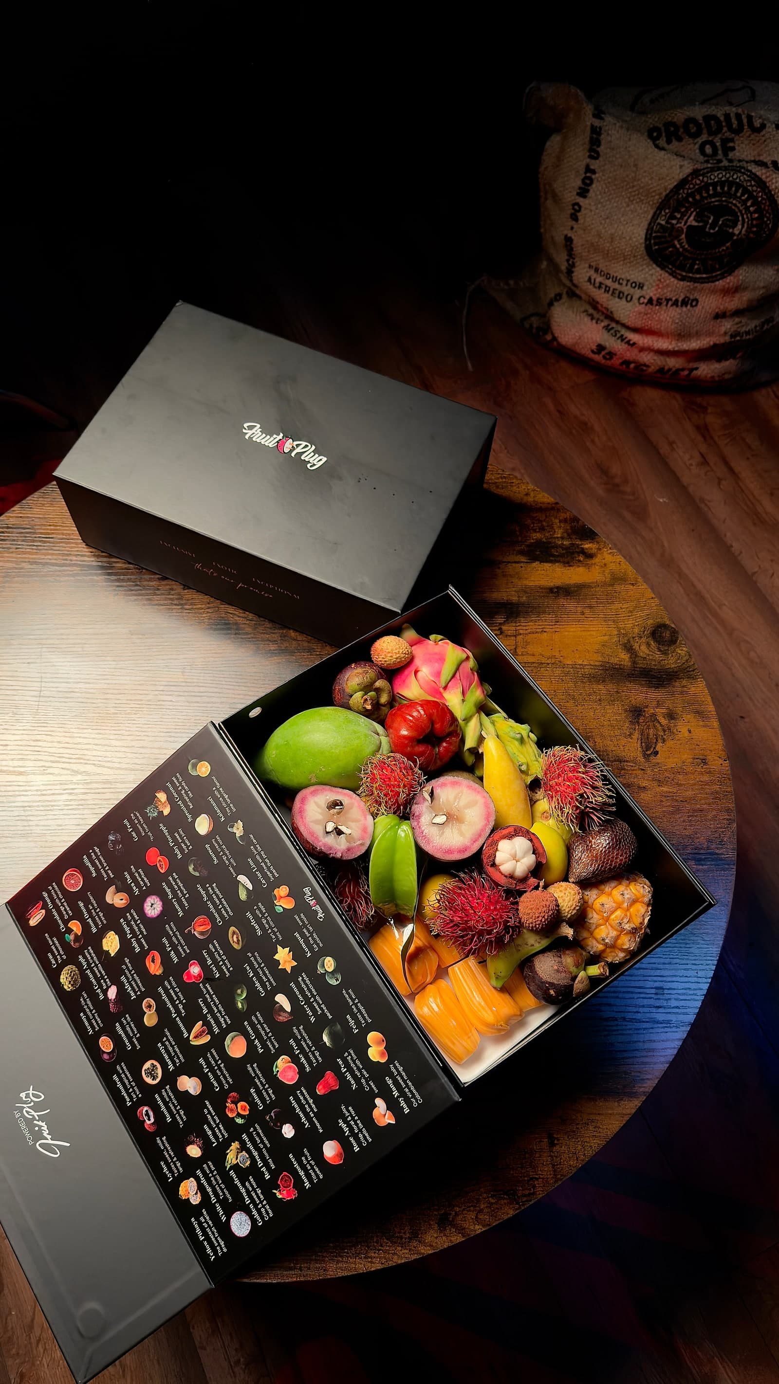 A Fruit Plug box opened on a wooden table, packed with mangosteen, dragon fruit, rambutan, papaya and banana, with the fruit-identification chart printed inside the lid.