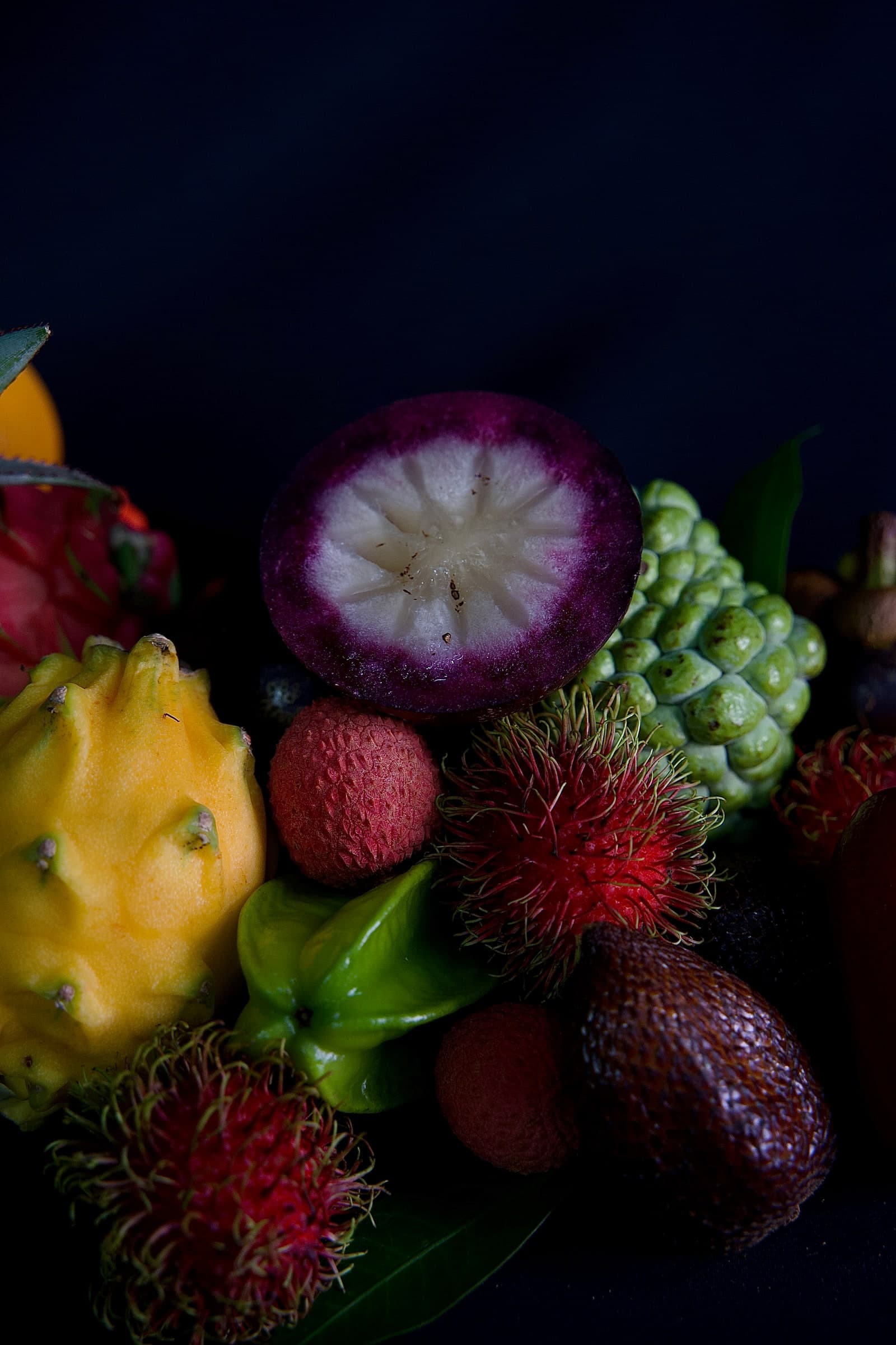 A close-up of an exotic fruit assortment — halved mangosteen showing white pith, yellow dragon fruit, rambutan, lychee, snake fruit, starfruit and custard apple, on a dark moody background.